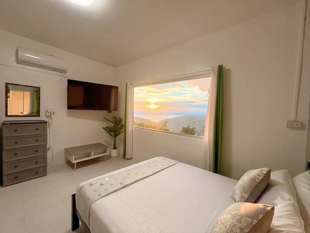 Een slaapkamer met een bed, een kaptafel, een aan de muur bevestigde televisie en een raam met uitzicht op de ondergaande zon.