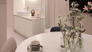 Una camera d'albergo moderna con un tavolo rotondo, uno specchio, un angolo cottura e un vaso decorativo con fiori.