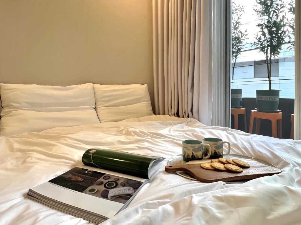 Uma cama bem arrumada com um travesseiro, um objeto cilíndrico verde, duas canecas e uma bandeja com biscoitos.