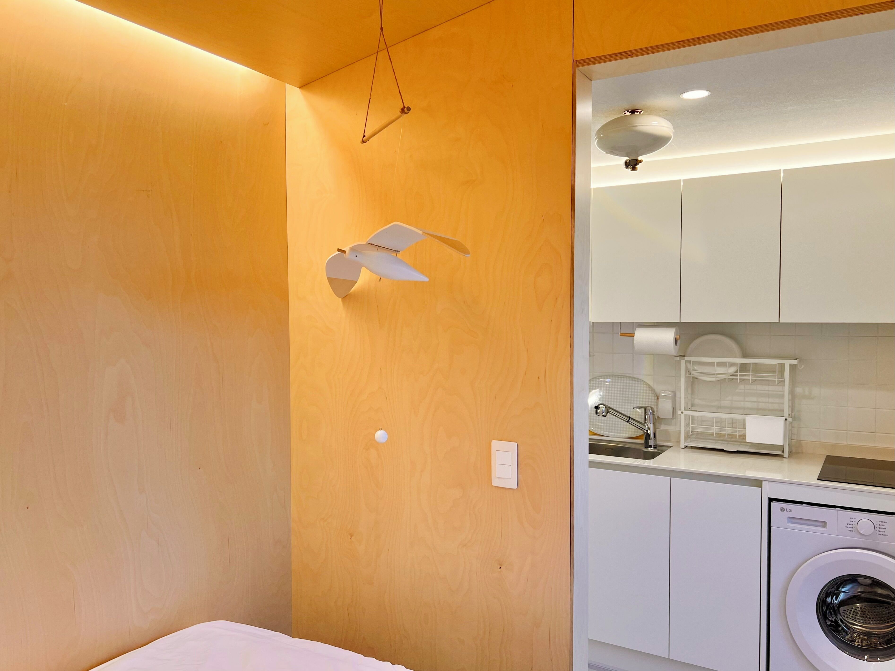 A compact room with a wooden wall, a ceiling fan, a washing machine, and a small kitchen area with a sink and a dish rack.