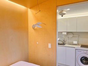 Habitación funcional con pared de madera, ventilador de techo, lavadora y una pequeña cocina con fregadero y escurreplatos.