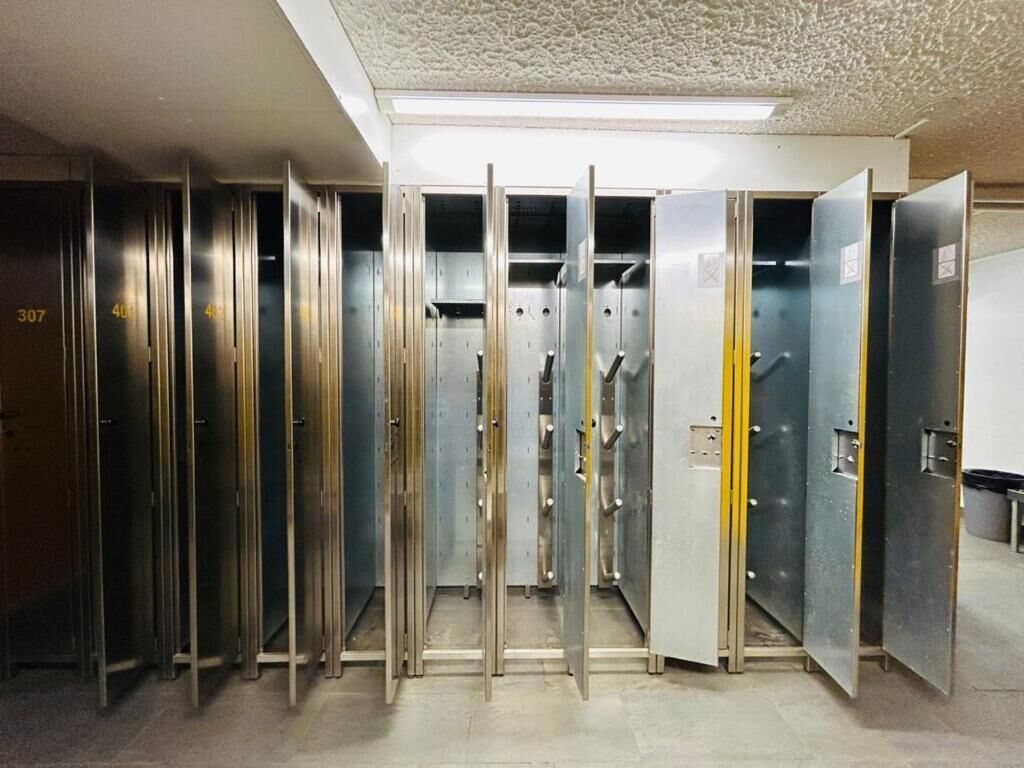 Une rangée de chambres d’hôtel métalliques, chacune portant un numéro, avec les portes ouvertes, laissant entrevoir les détails de l’intérieur.