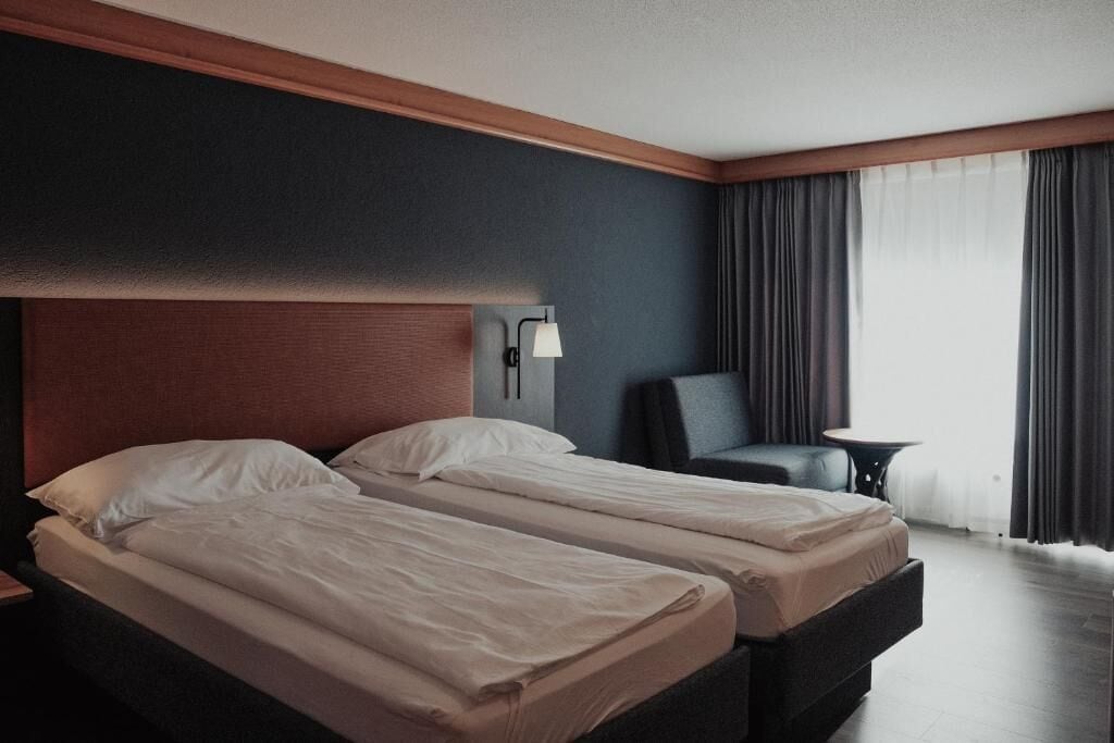 Quarto de hotel com duas camas, uma mesinha de cabeceira, uma cadeira, uma pequena mesa e uma janela com cortinas.