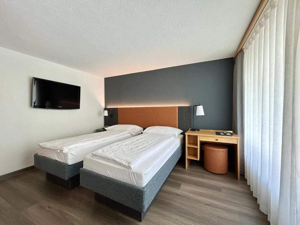 A hotel room with two beds, a desk, a chair, a television, and a window with curtains.