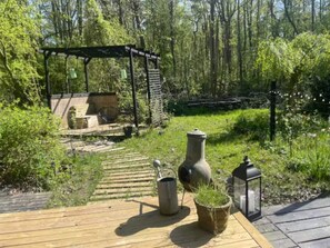 Una terraza de madera con un área de estar rústica al aire libre, una pérgola y un sendero de jardín que atraviesa un área boscosa.