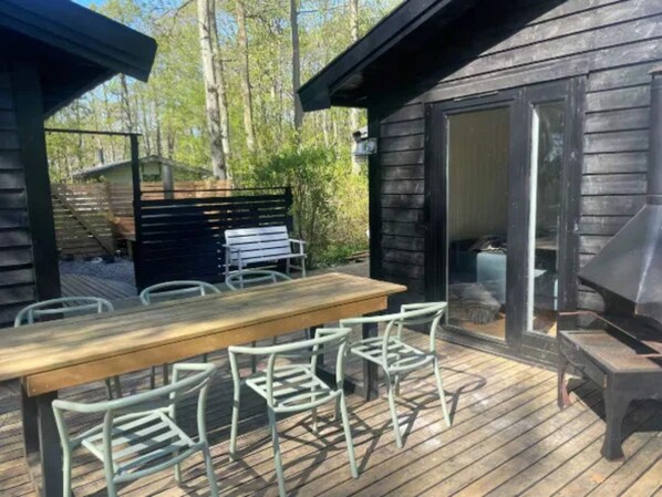 Una terraza de madera con una mesa y sillas, una parrilla y un ventanal corredizo que da a una zona cubierta.