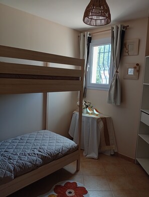 Ett rum med en våningssäng, ett litet bord med en lampa, ett fönster med gardiner och en hylla som är monterad på väggen.