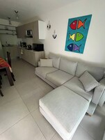Una sala moderna con un sofá seccional gris, un comedor con mesa y sillas, y una cocina al fondo.