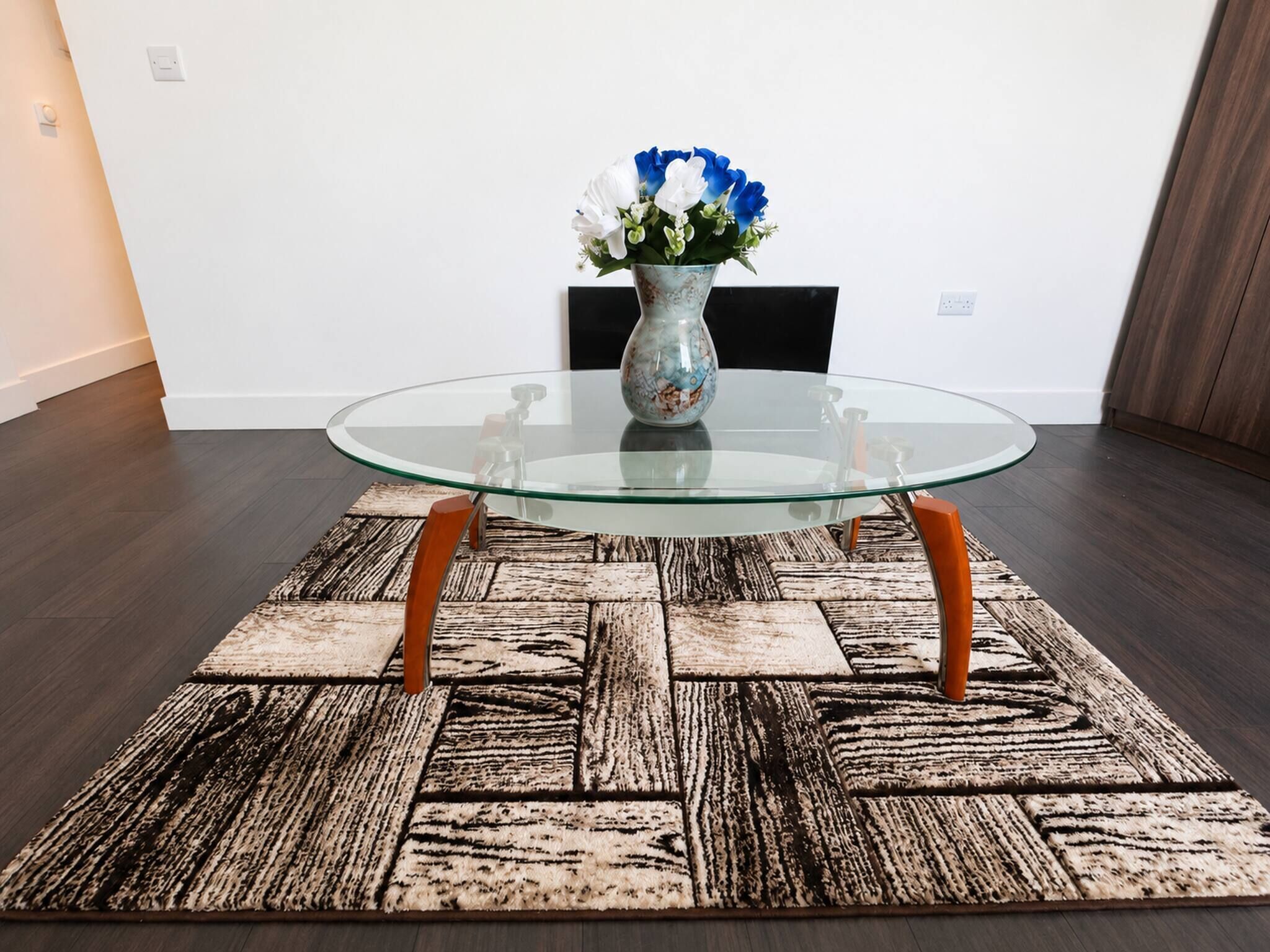 Une table avec un plateau en verre et des pieds en bois, un tapis à motifs et un vase rempli de fleurs placé sur un mur uni.