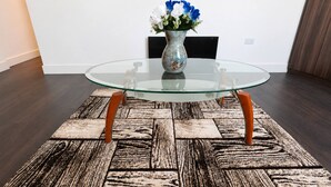 Uma mesa com tampo de vidro e pés de madeira, um tapete estampado e um vaso com flores em uma parede lisa.