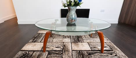 A glass-top table with wooden legs, a patterned rug, and a vase with flowers on a plain wall.