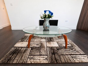 Ein Tisch mit Glasplatte und Holzbeinen, ein gemustertes Teppich und eine Vase mit Blumen an einer schlichten Wand.