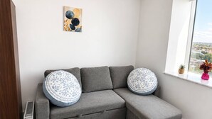 Um sofá modular cinzento com almofadas estampadas, um radiador, uma janela com vista e uma obra de arte abstrata na parede.