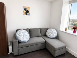 Ein graues Ecksofa mit gemusterten Kissen, eine Heizung, ein Fenster mit Blick und ein abstraktes Kunstwerk an der Wand.