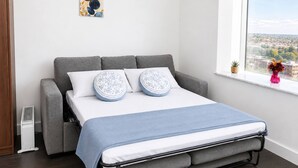 Quarto moderno com sofá cinza, cama com manta azul, almofadas estampadas e vista para o horizonte urbano pela janela.