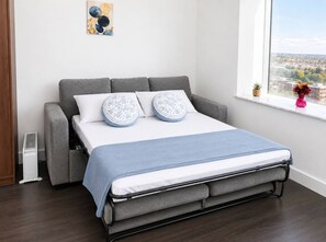 Ein modernes Schlafzimmer mit grauem Sofa, Bett mit blauer Decke, gemusterten Kissen und Blick auf eine Stadtlandschaft durch ein Fenster.