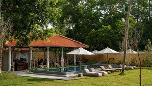 A poolside area with lounge chairs and umbrellas, a covered seating area, and a building with a tiled roof. - Rekawa Wellness Resort  (Netolpitiya)