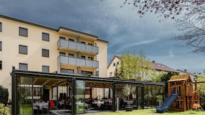 Un edificio moderno con un espacio exterior cubierto de vidrio para comer y una estructura de juegos para niños.