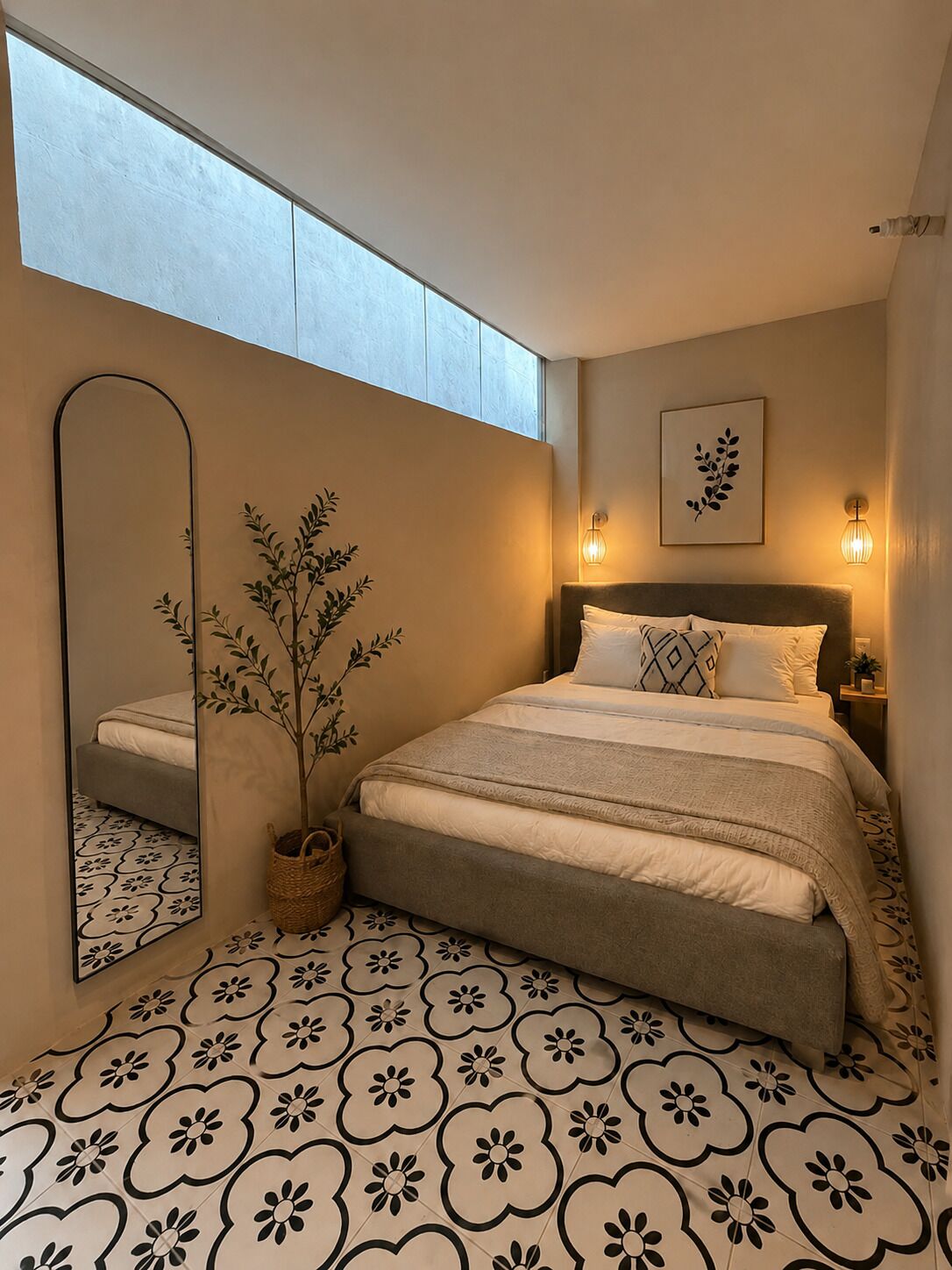 A bedroom with a bed, a mirror, a potted plant, a wall-mounted light, and a framed artwork.
