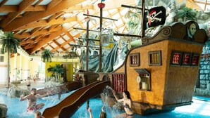 Parque acuático interior con zona de juegos temática de piratas, que incluye un tobogán y una estructura con forma de barco.