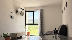 A modern room with a glass table, a sofa, a TV mounted on the wall, and a window with a view of greenery.