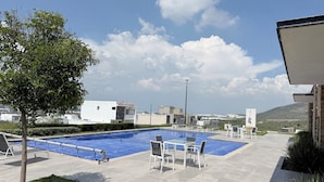 A swimming pool with lounge chairs and a tree, surrounded by buildings and a clear sky.