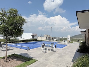 A swimming pool with lounge chairs and a tree, surrounded by buildings and a clear sky.