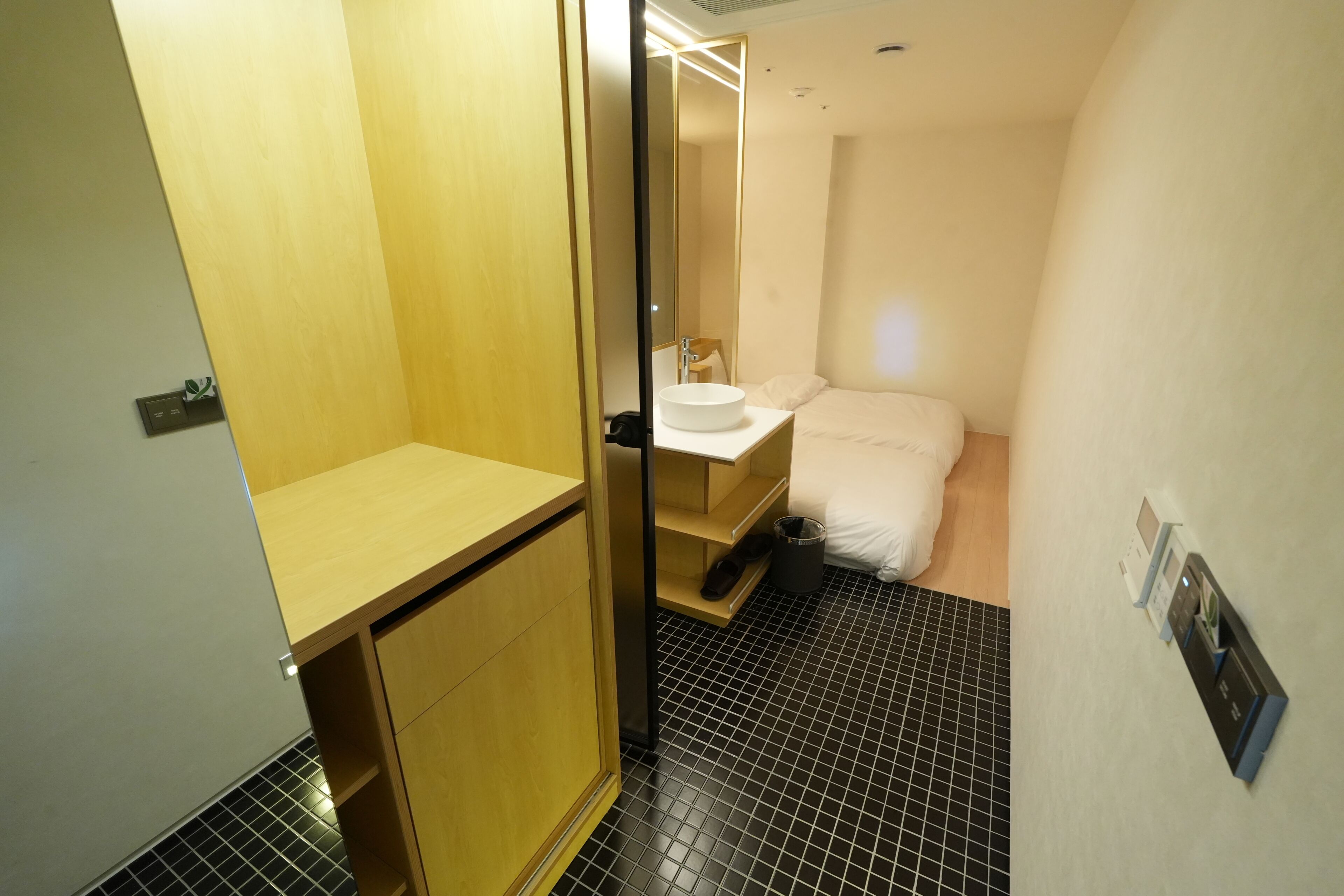 A hotel room with a wooden wardrobe, a bedside table, a white sink, and a black tiled floor.