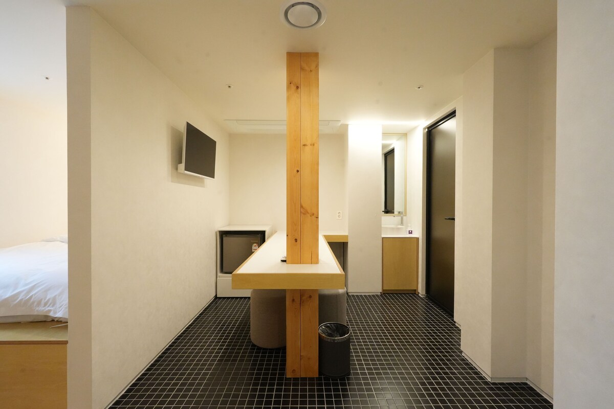 A hotel room with a bed, a small desk, a trash bin, and a kitchenette.
