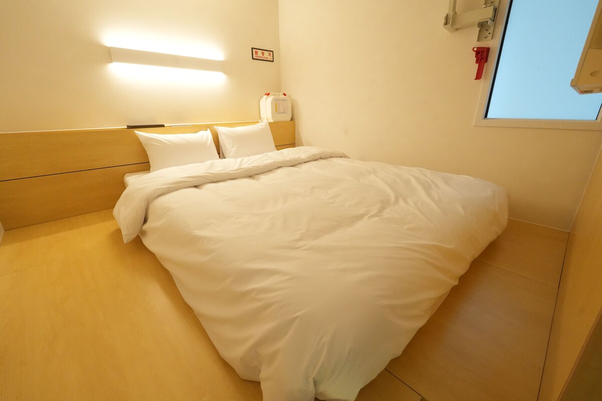 A hotel room with a bed, a nightstand, a wall-mounted light, and a window.