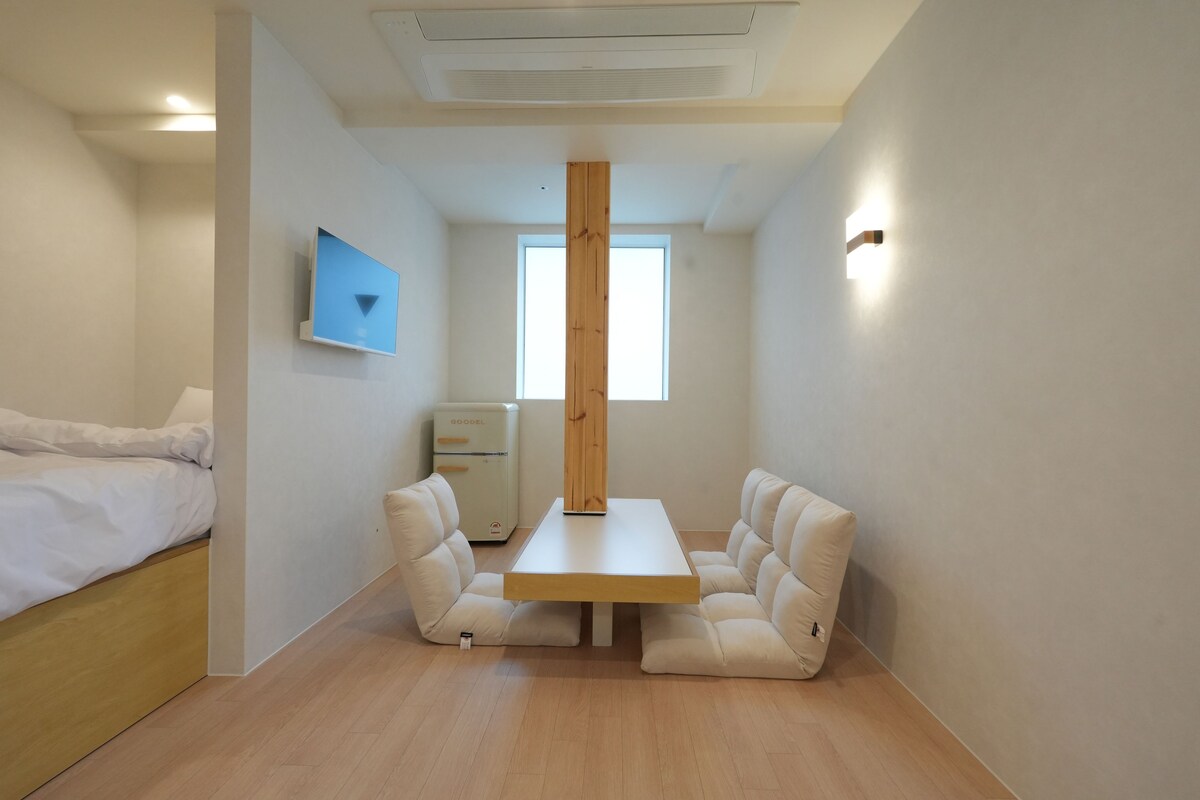 A compact room with a bed, a small kitchenette, a wooden table with chairs, and a window providing natural light.