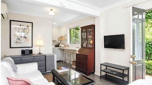 A modern living room with a sofa, coffee table, and a TV. There is a kitchen area with a dining table and chairs. The room has a large window and a door leading outside. - “Highly rated Blackheath garden studio — 4.94 from over 1,000 Airbnb guests” (Blackheath)