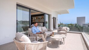 A man seated on a balcony with a laptop, surrounded by wicker furniture and a glass railing overlooking a coastal view. - VILLA STATUS BY VACATION MARBELLA (Estepona)
