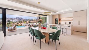 A modern kitchen with a central island, a dining area with green chairs, and a view of a pool and landscape. - VILLA STATUS BY VACATION MARBELLA (Estepona)