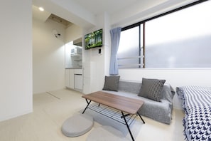 Un espace de vie compact comprenant un coin cuisine, un canapé agrémenté de coussins à motifs, une table en bois et une fenêtre avec des rideaux.