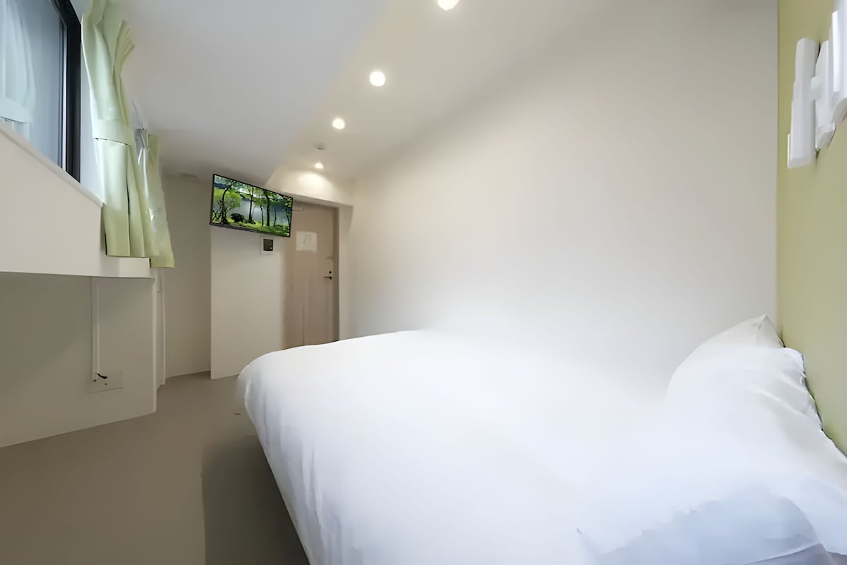 Une chambre d’hôtel avec un lit, une fenêtre avec des rideaux, un téléviseur fixé au mur et une porte.