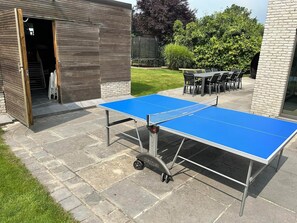 An outdoor ping pong table set up on a patio with a stone floor, surrounded by a brick wall and a wooden structure with an open door.