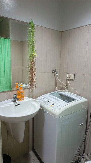 A bathroom with a white sink, a mirror, a green curtain, a white washing machine, and a hanging plant. - Enjoyable 1-bedroom condo with AC in Pasay City (Pasay City)