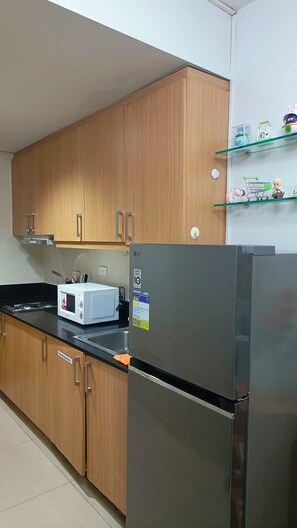 A kitchen with wooden cabinets, a black refrigerator, a white microwave, and a glass shelf with decorative items. - Enjoyable 1-bedroom condo with AC in Pasay City (Pasay City)