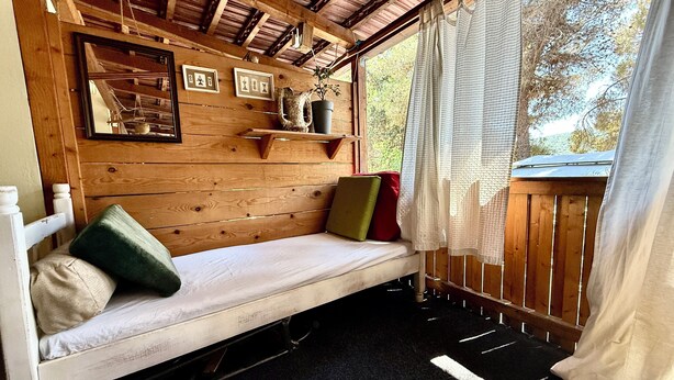Interior de una cabaña de madera con una cama, almohadas, un estante con adornos y una ventana con cortinas.