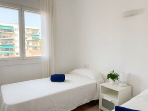 Ein Zimmer mit einem Einzelbett, einer weißen Bettdecke, einem blauen Kissen, einem Nachttisch mit Pflanze und einem Fenster mit Blick auf Gebäude.
