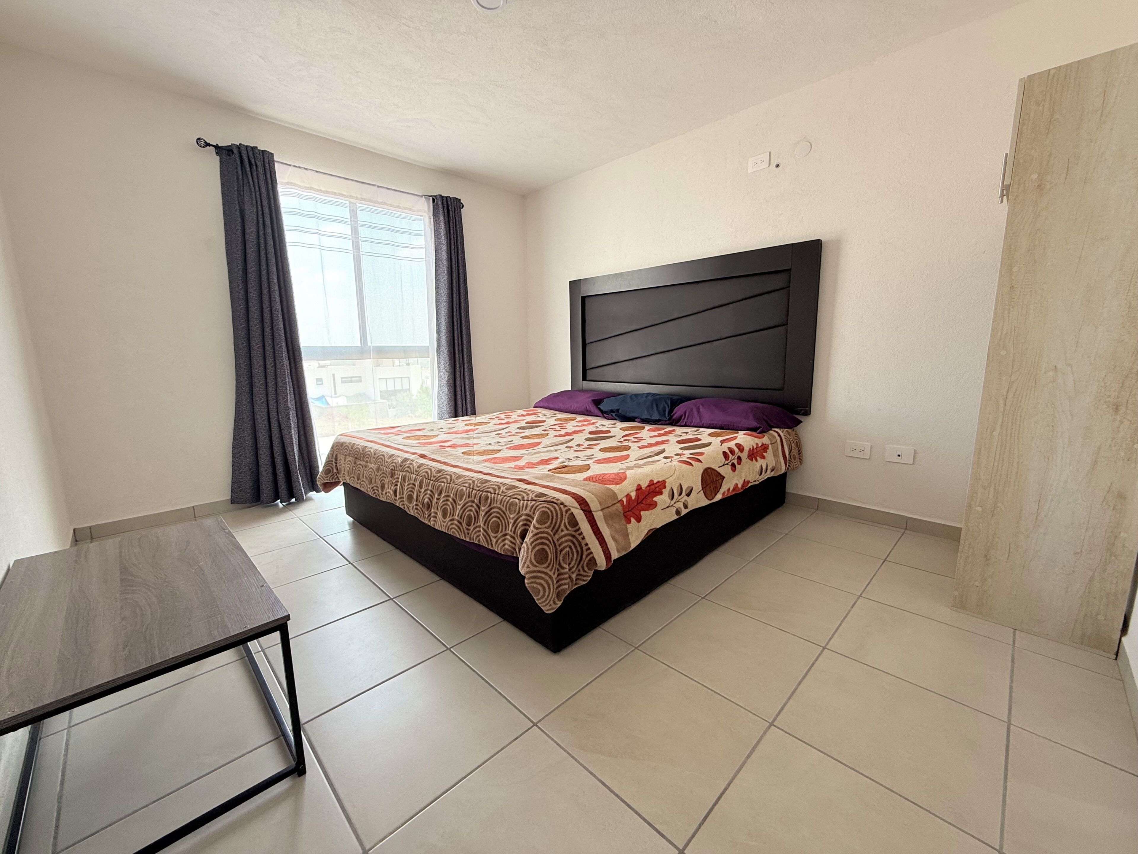 Un dormitorio con cama, mesita de noche, ventana con cortinas y vistas al exterior.