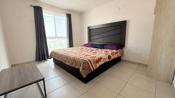 Un dormitorio con cama, mesita de noche, ventana con cortinas y vistas al exterior.