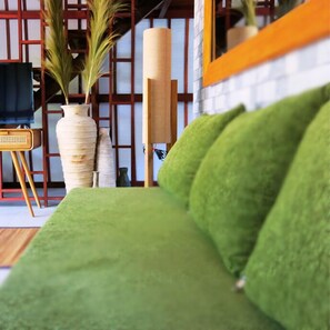 A green sofa in a room with a wooden chair, a tall vase with plants, and a large mirror on the wall.