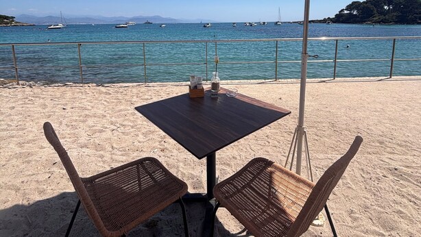 Ein Tisch am Strand mit zwei Korbstühlen und einem großen Sonnenschirm, mit Blick auf ein ruhiges Meer mit Booten.