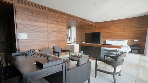 A modern hotel room with a wooden panel wall, a flat-screen TV, a desk with a chair, a sofa, and a bed. - The Lalu Qingdao (Qingdao)