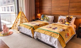 Habitación de hotel con dos camas, cabecera de madera, mesita de noche, lámpara, un oso de peluche, un caballo de juguete y una pequeña tienda de campaña.