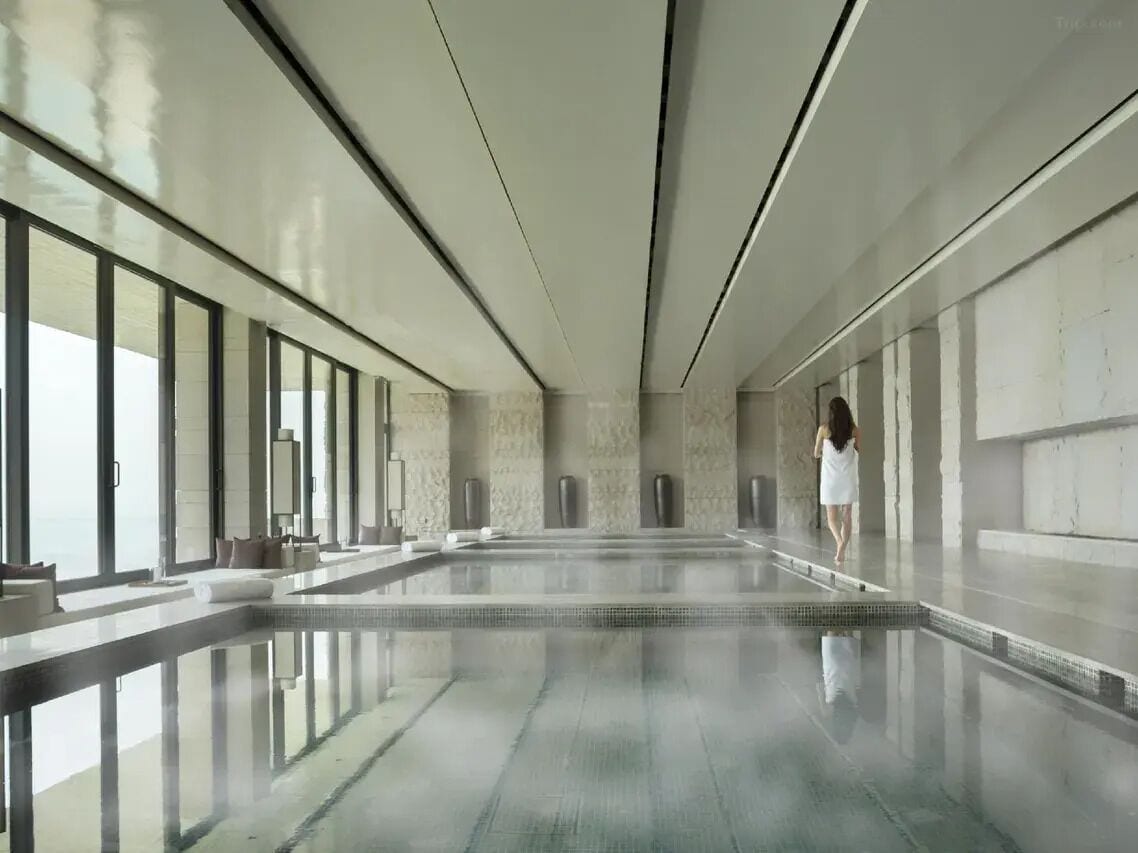 A spacious indoor swimming pool with clear glass walls, a modern design, and a person walking by.