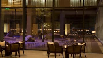 A dining area with tables and chairs, a large window with a view of a landscaped area and city lights.