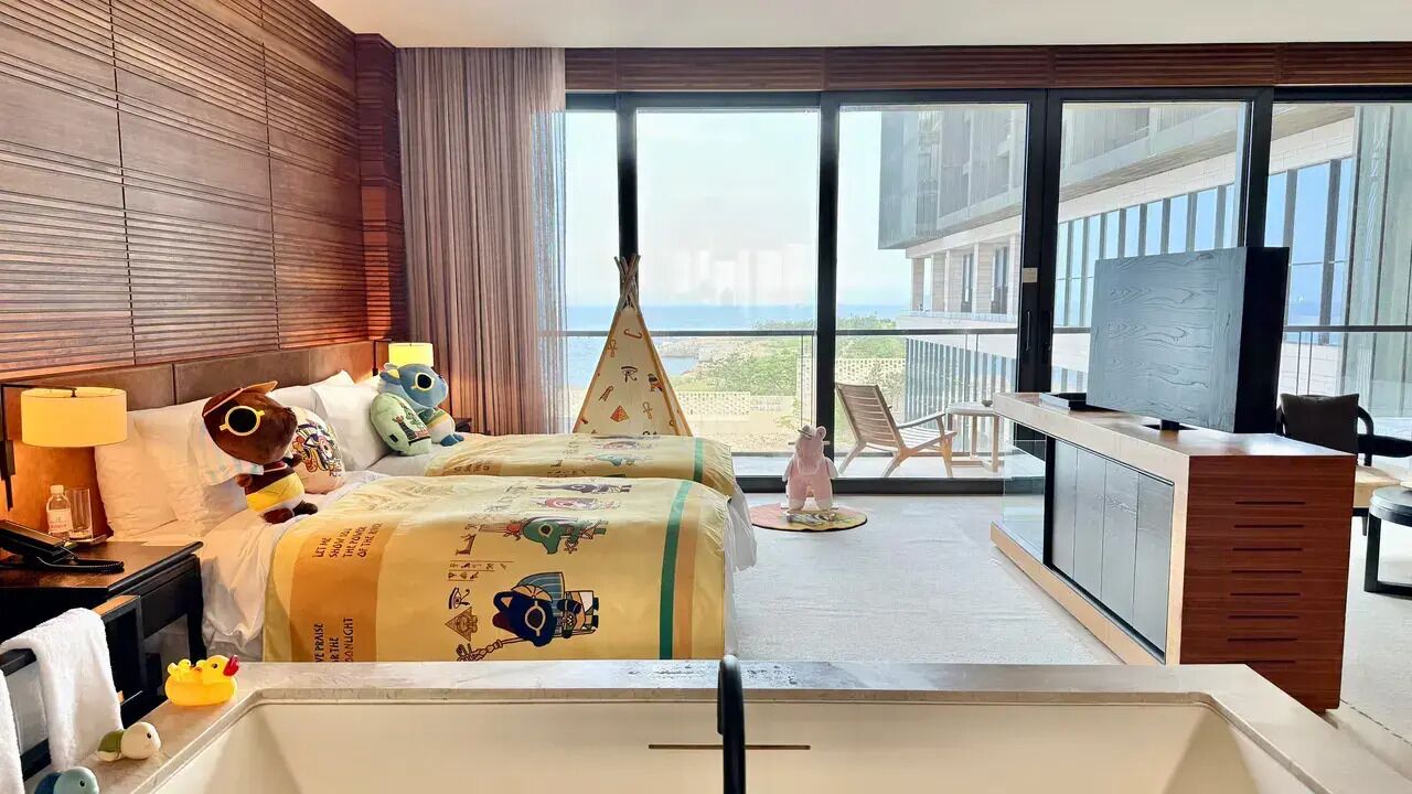 A hotel room with a large bed, a desk, a chair, a television, and a view of the ocean through large windows.